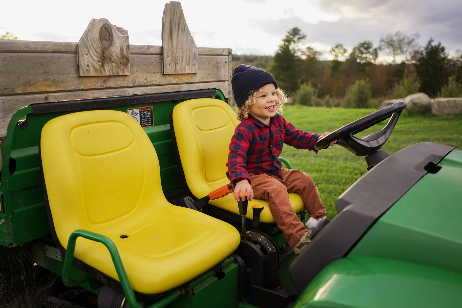 Child in a blue beanie and red plaid shirt steering a green tractor with yellow seat