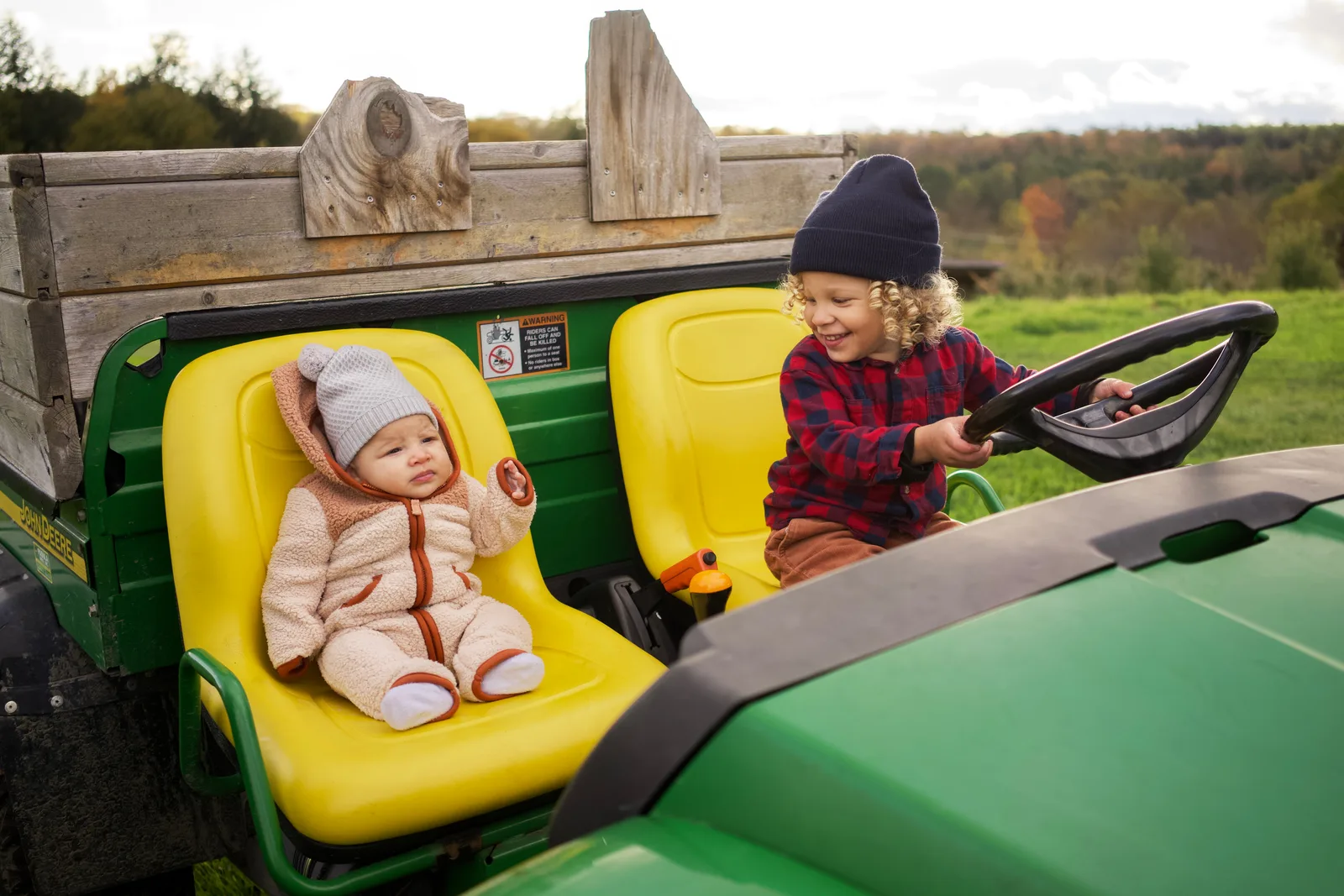 Baby in a light gray knit hat and cream jacket with orange trim seated in a yellow tractor seat