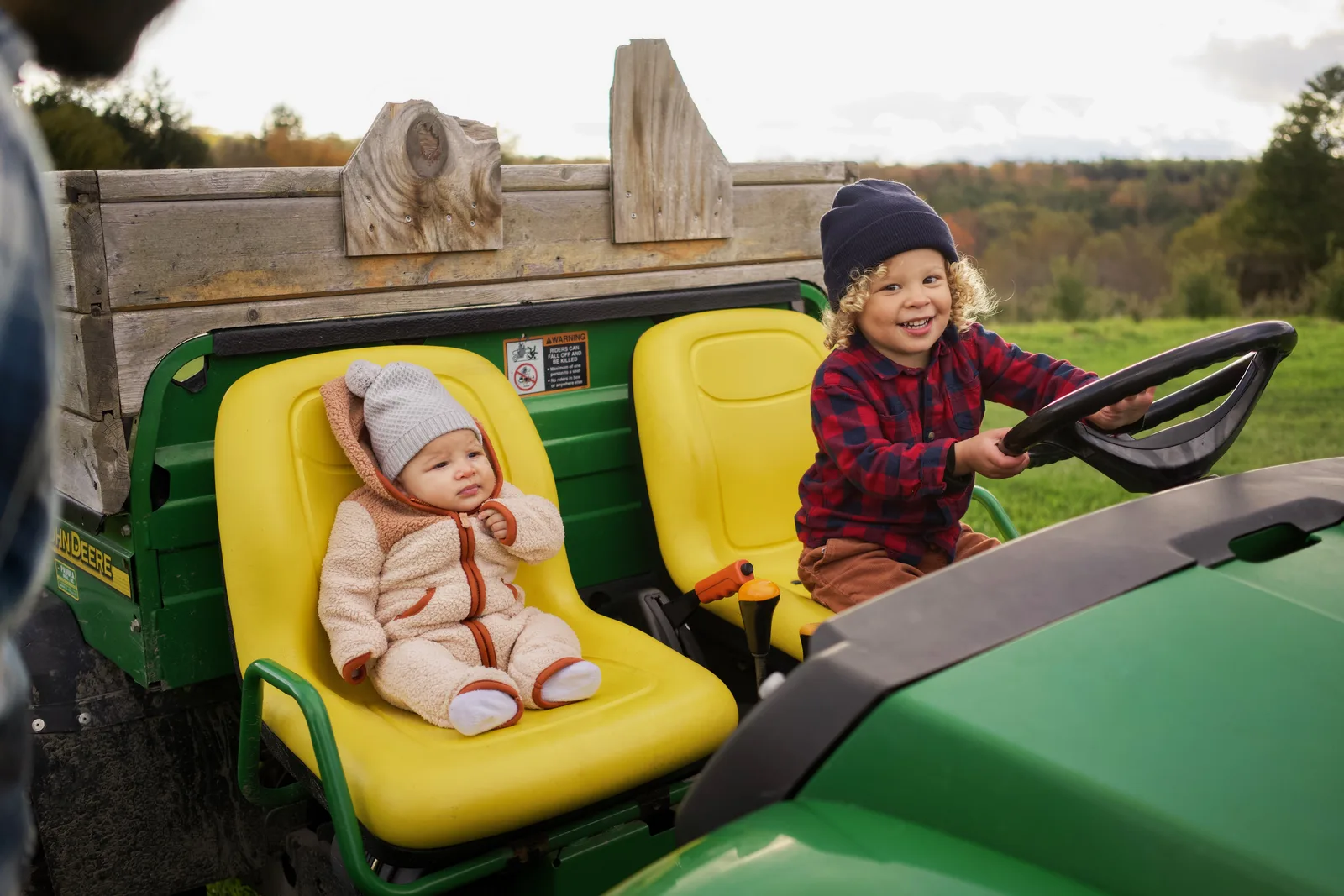 Two children in a green tractor, one in a cream bear suit on a yellow seat, the other steering