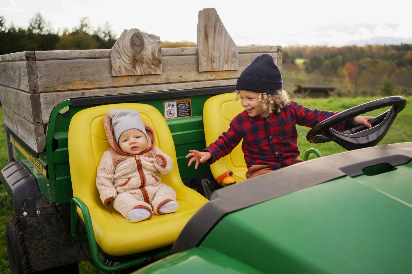 Child in red plaid shirt and blue beanie steering green tractor, baby in cream bear suit seated
