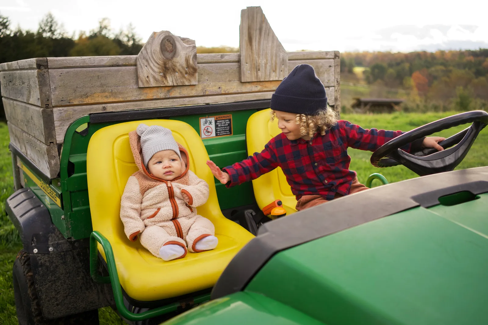 Child in red plaid shirt and blue beanie reaching toward baby in cream bear suit on tractor