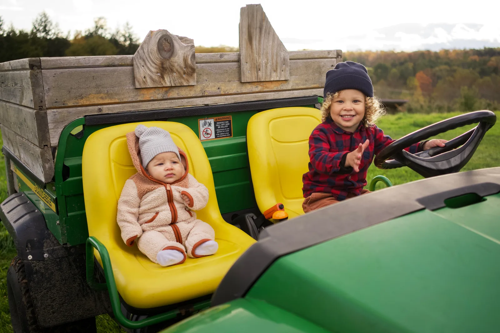 Child in red plaid shirt and blue beanie pointing at baby in cream bear suit on tractor