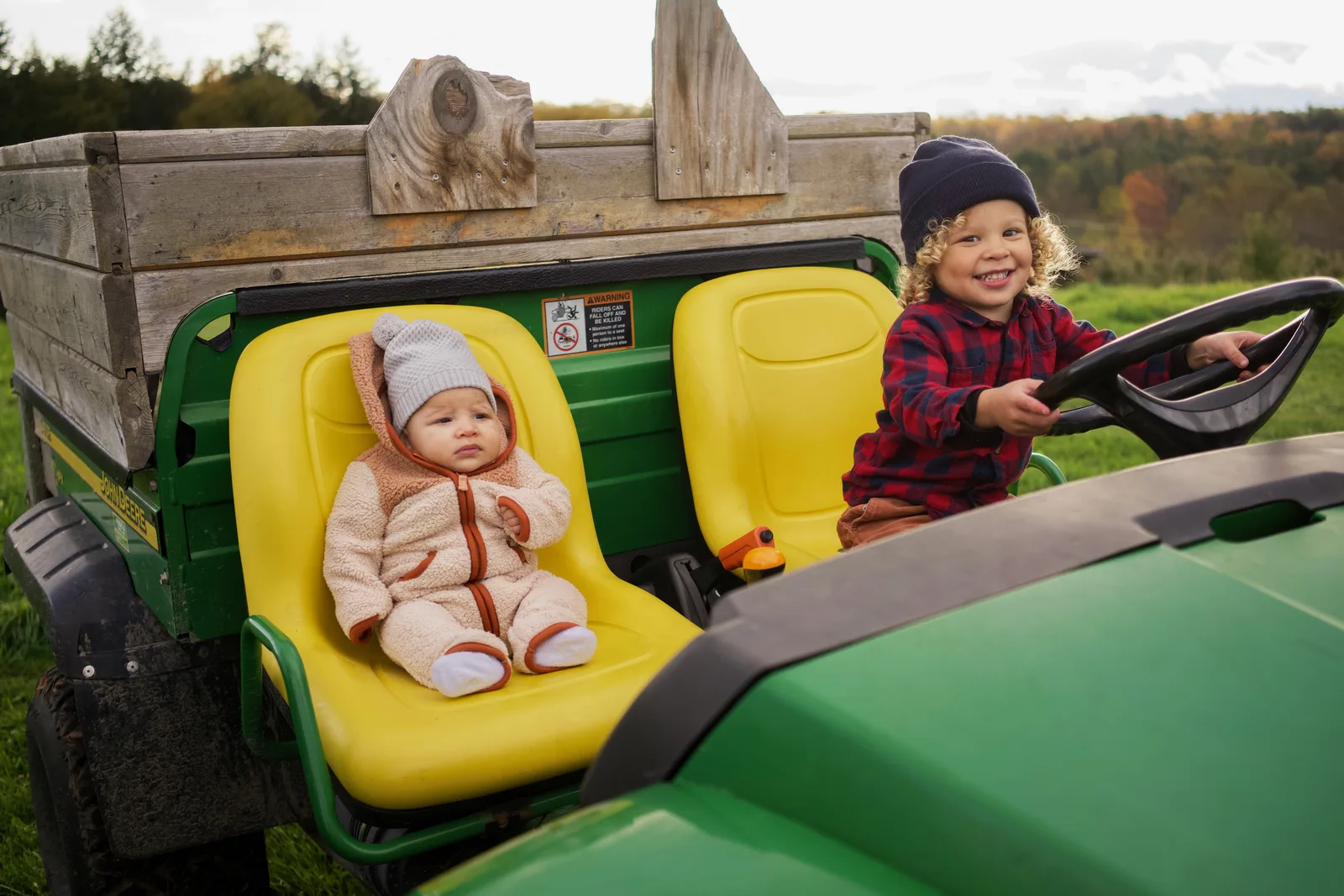 Child in red plaid shirt and blue beanie holding steering wheel, baby in cream bear suit seated
