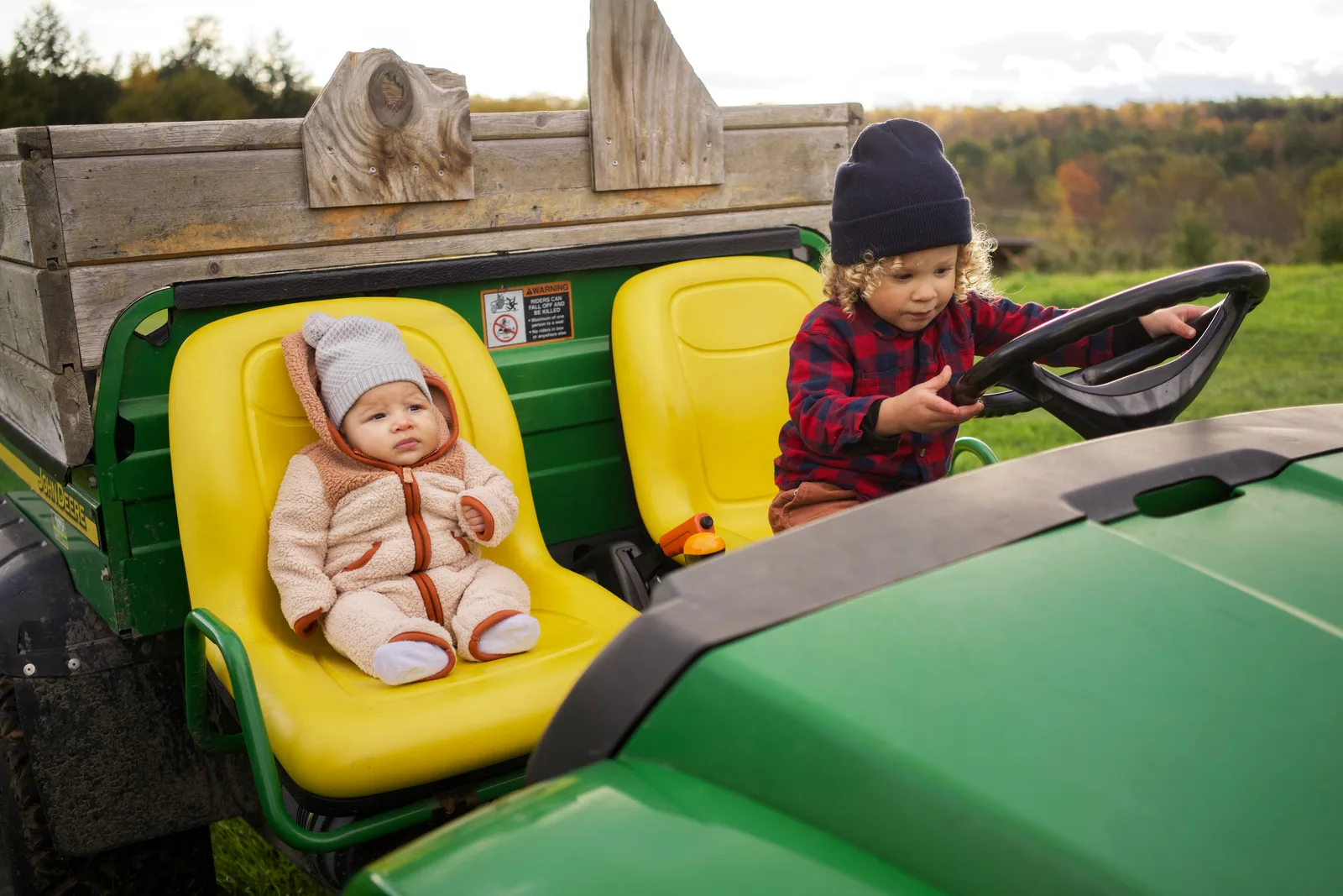 Child in red plaid shirt and blue beanie gripping steering wheel, baby in cream bear suit seated