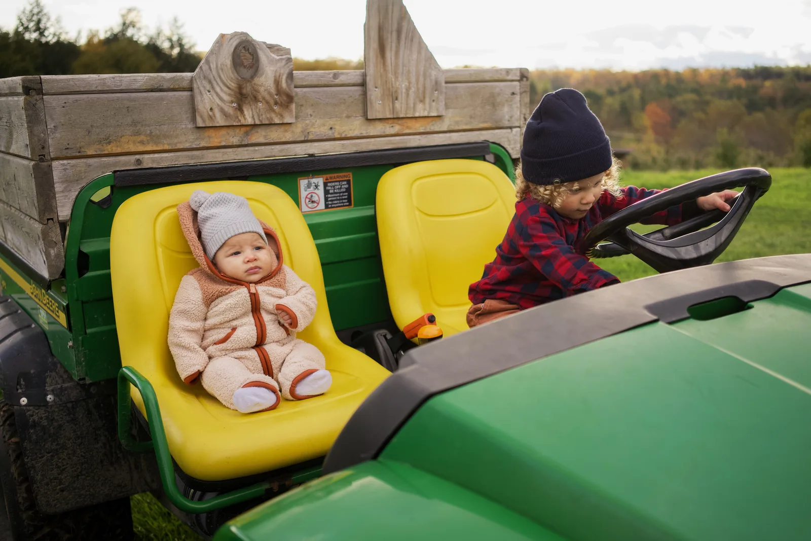 Child in red plaid shirt and blue beanie turning steering wheel, baby in cream bear suit seated