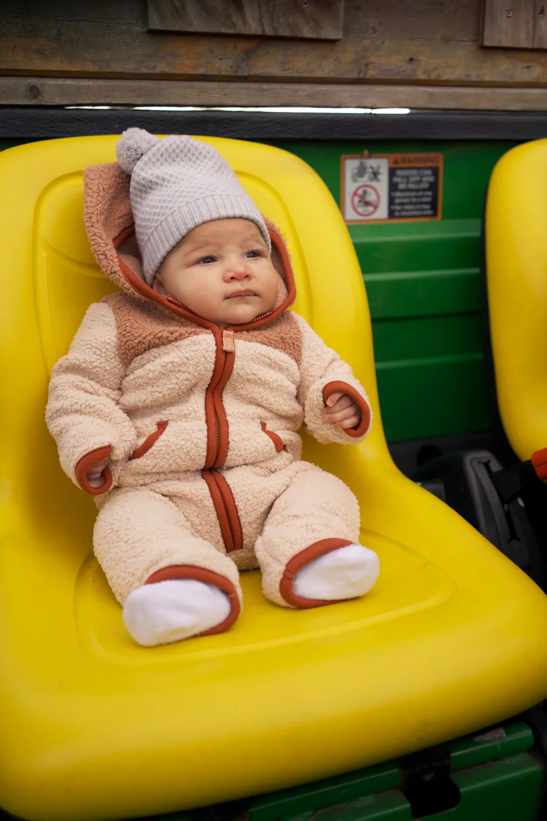 Baby in cream bear suit and gray knit hat seated on a yellow tractor seat