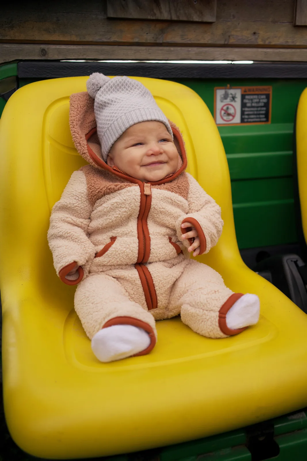 Baby in cream bear suit with gray knit hat seated on yellow tractor seat