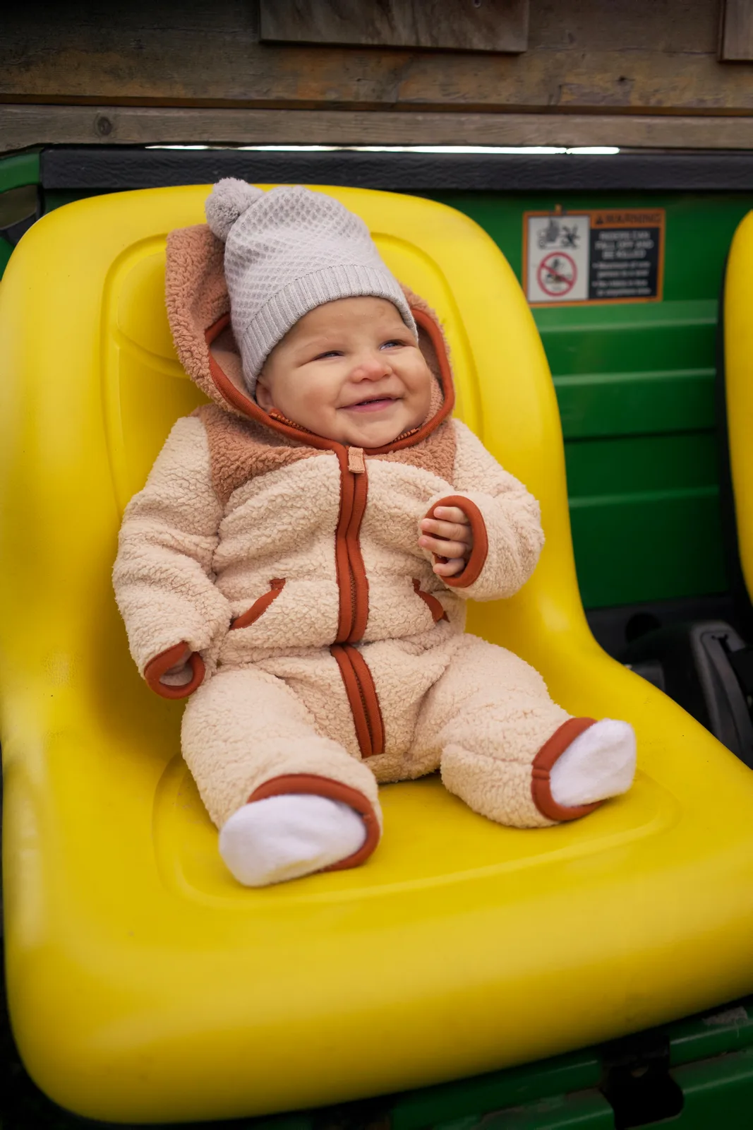 Baby in cream bear suit with gray knit hat sitting on yellow tractor seat, green tractor behind