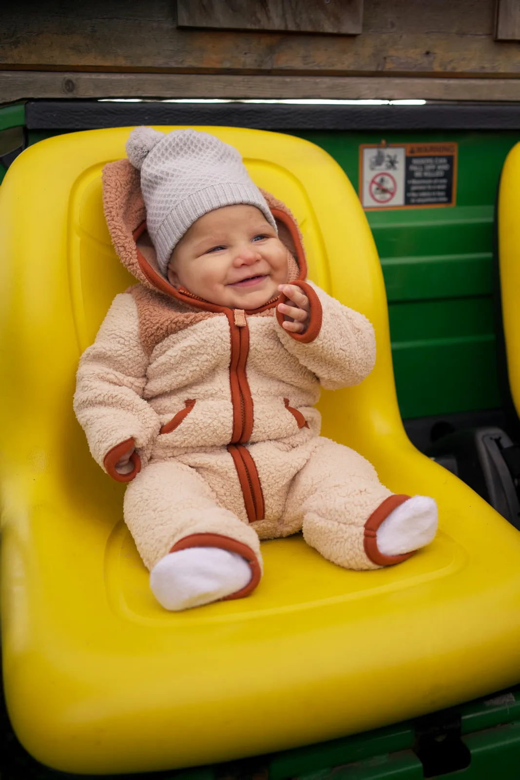 Baby in cream bear suit and gray hat on yellow seat, green tractor with warning sign