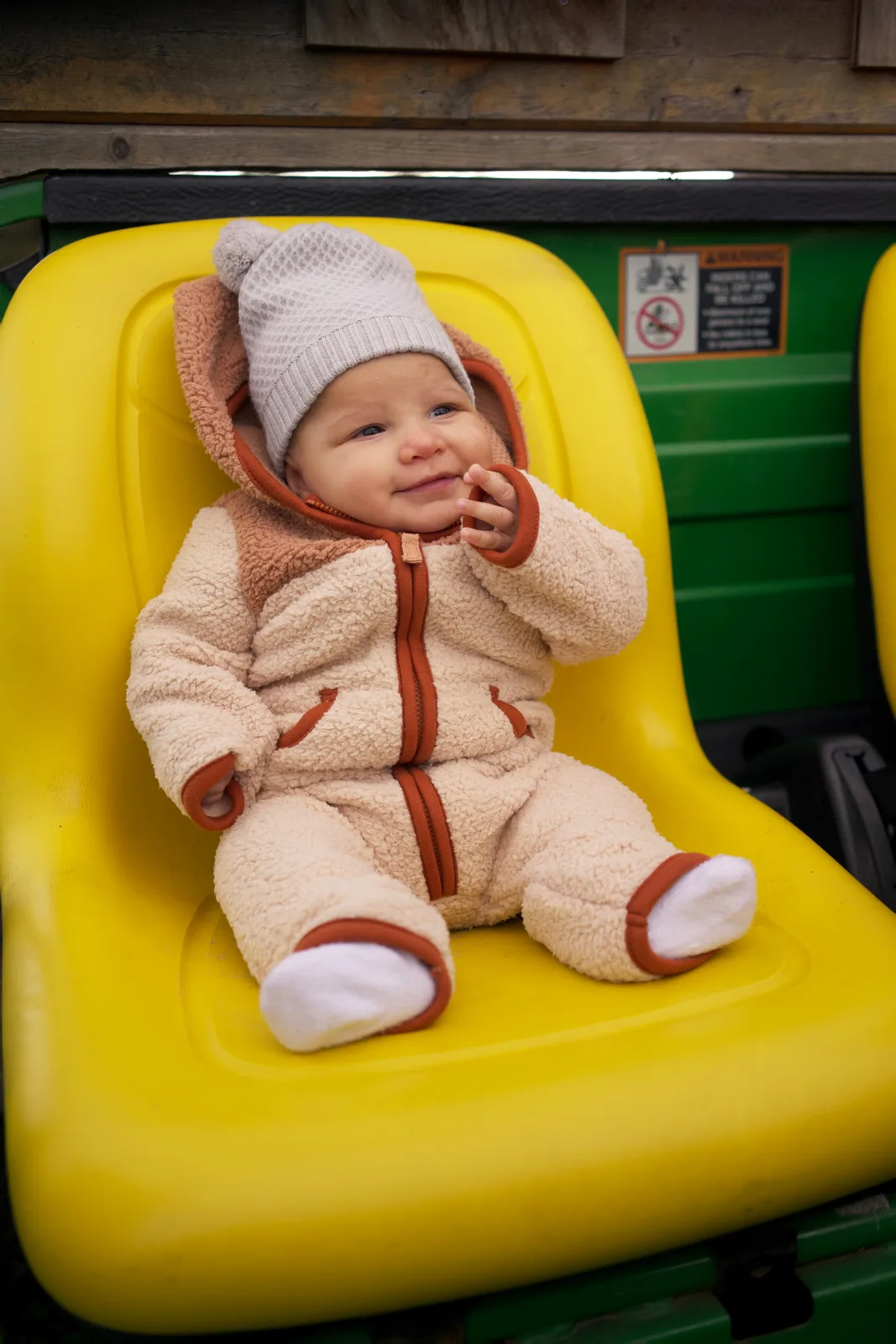 Baby in cream bear suit and gray beanie on yellow seat, green tractor interior visible