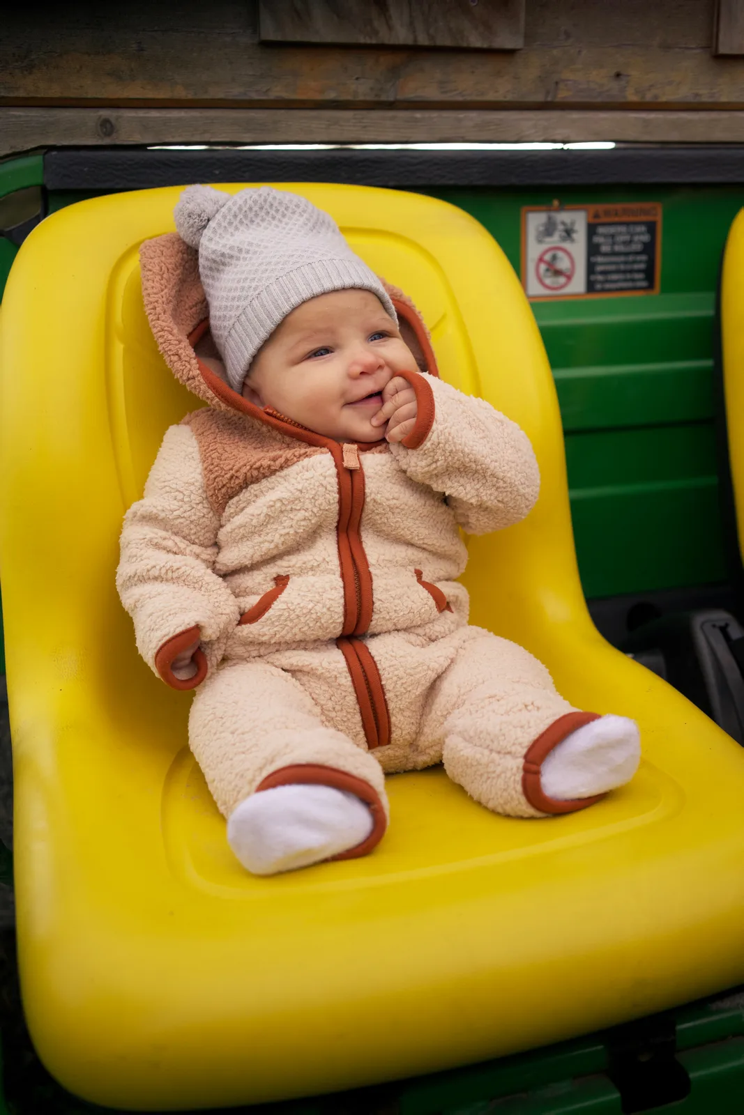 Baby in cream bear suit and gray beanie on yellow seat, green tractor with safety label