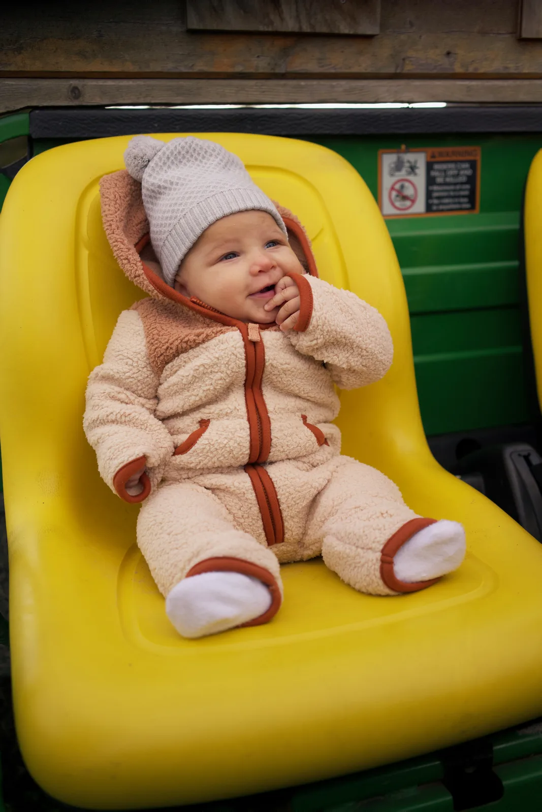 Baby in cream bear suit and gray hat, yellow seat, green tractor with warning sign