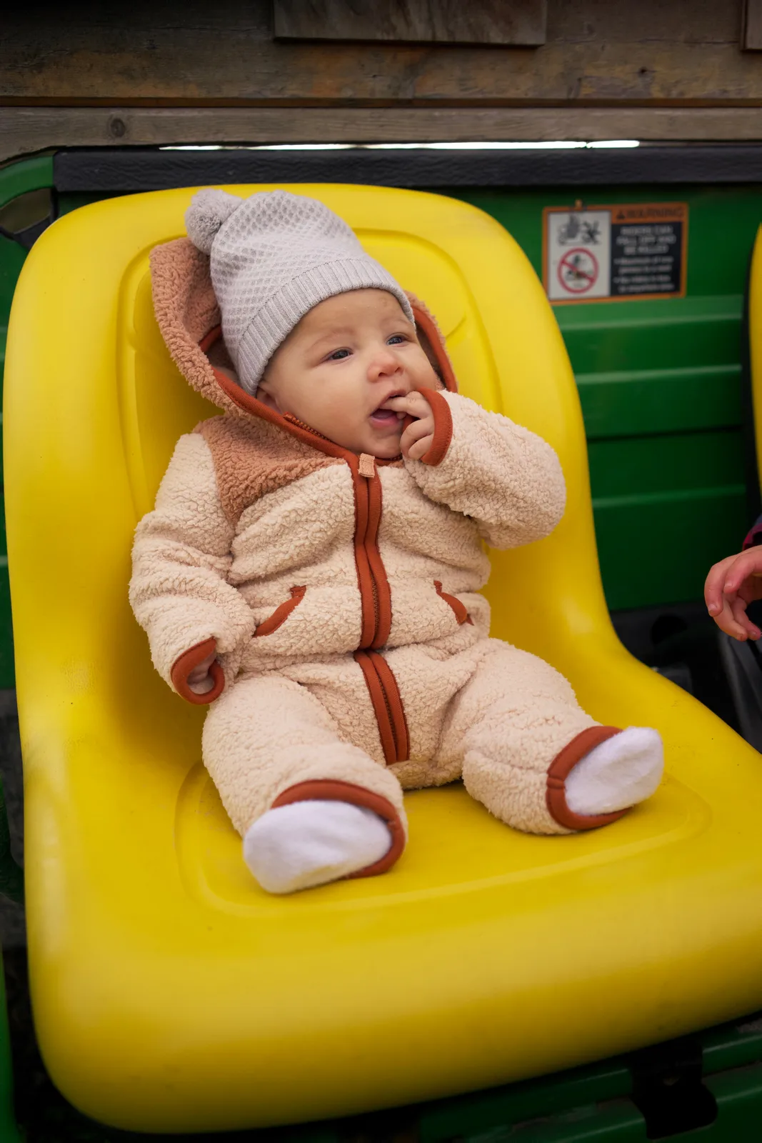 Baby in cream bear suit and gray hat on yellow seat, green tractor with partial hand visible