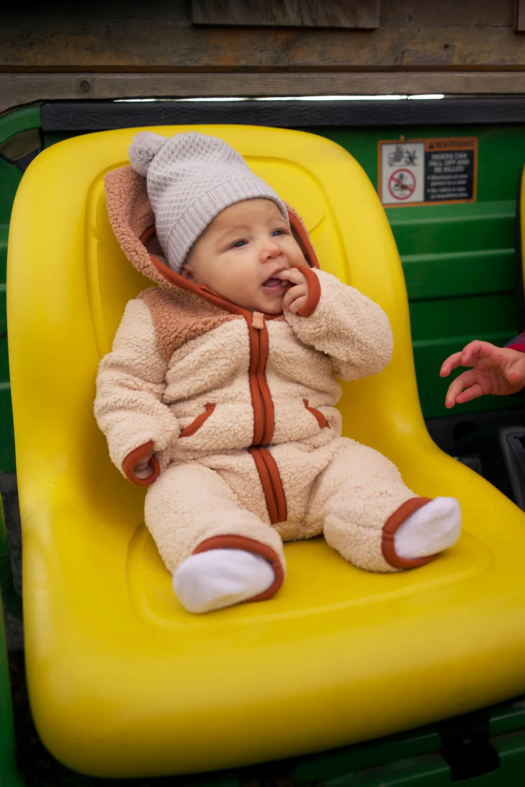 Baby in cream bear suit with gray knit hat on yellow tractor seat, green tractor behind