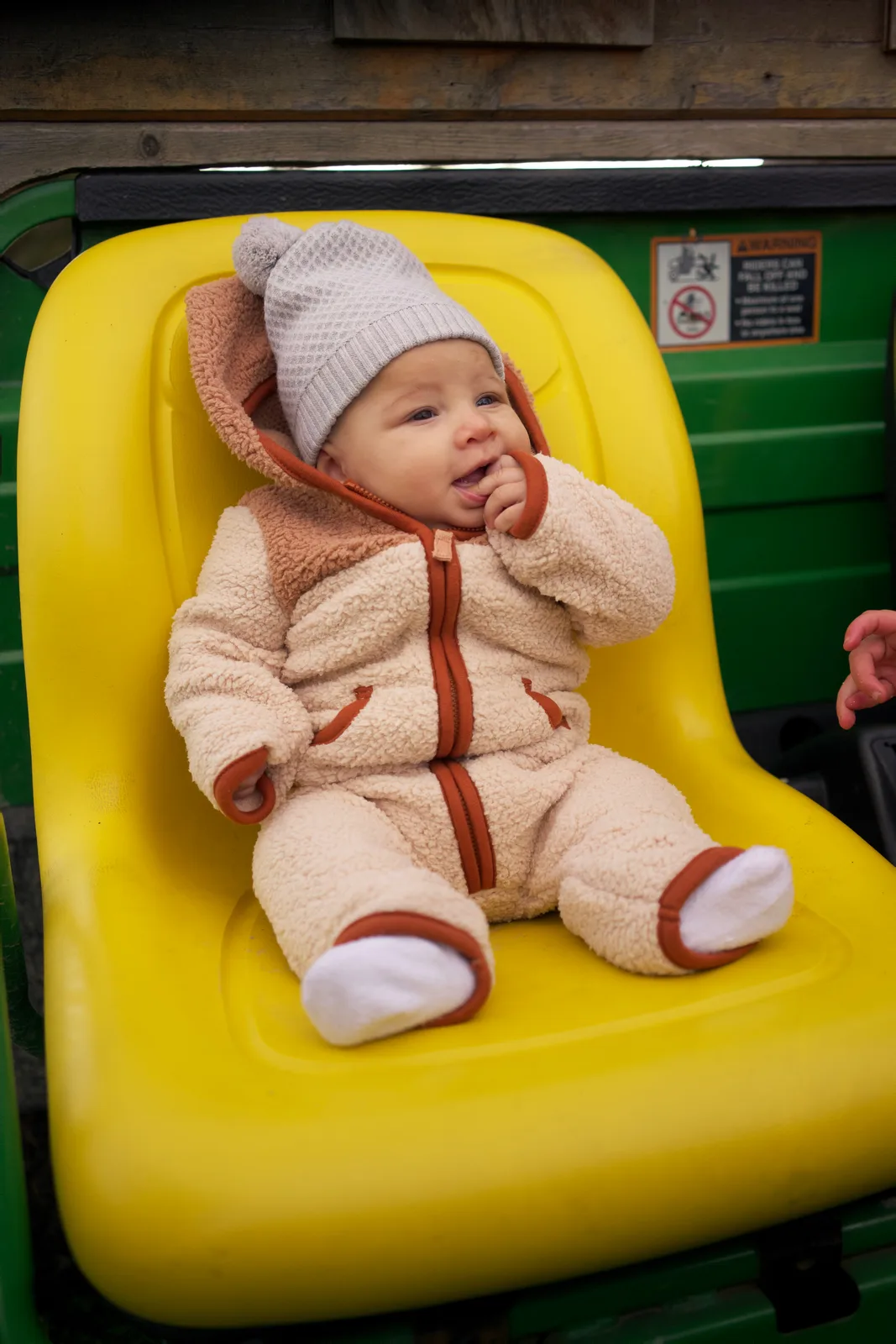 Baby in cream bear suit and gray hat on yellow seat, green tractor with warning sign