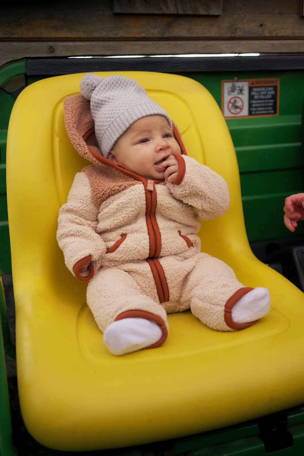 Baby in cream bear suit and gray beanie on yellow seat, green tractor interior visible