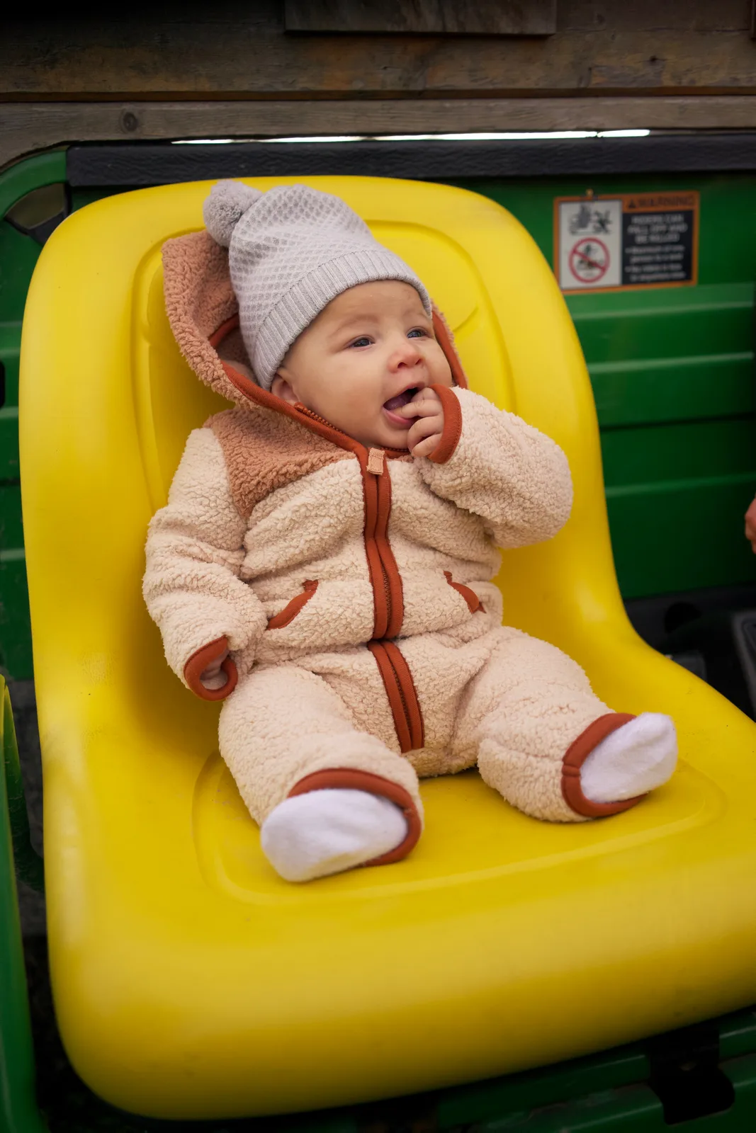 Baby in cream bear suit with gray hat, yellow seat, green tractor and wooden wall