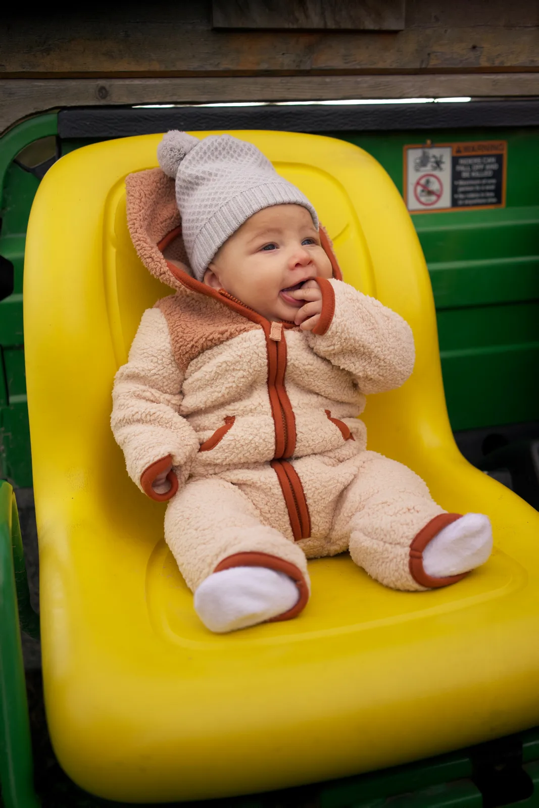 Baby in cream bear suit and gray beanie on yellow seat, green tractor with safety label