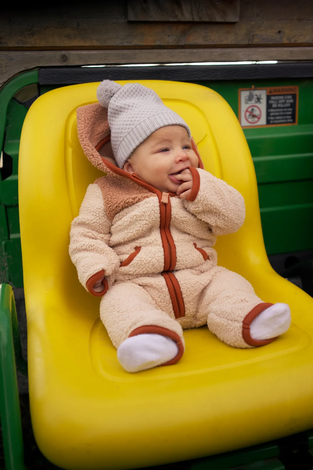 Baby in cream bear suit and gray hat on yellow seat, green tractor with partial hand visible
