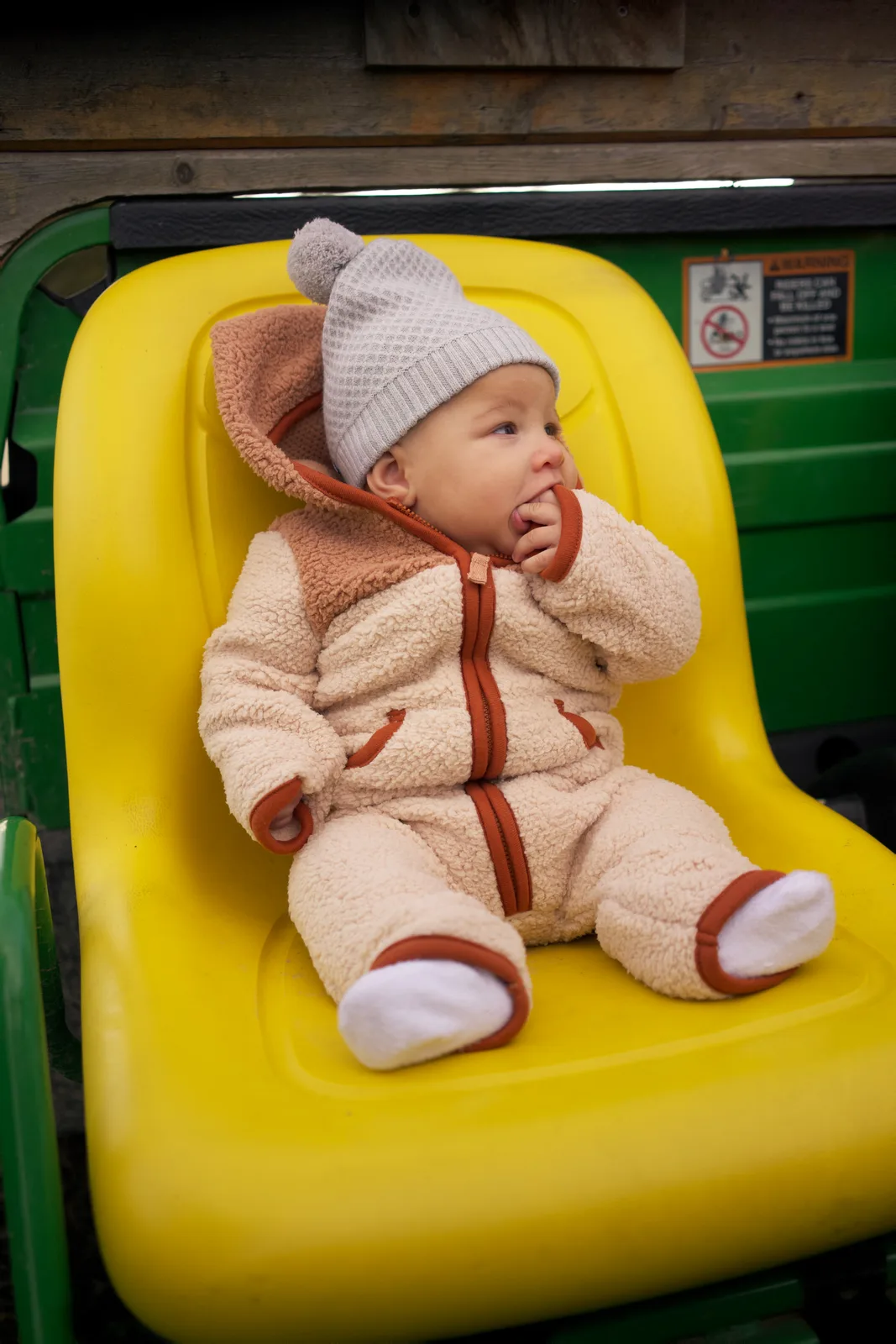 Baby in cream bear suit and gray beanie on yellow seat, green tractor and wooden wall