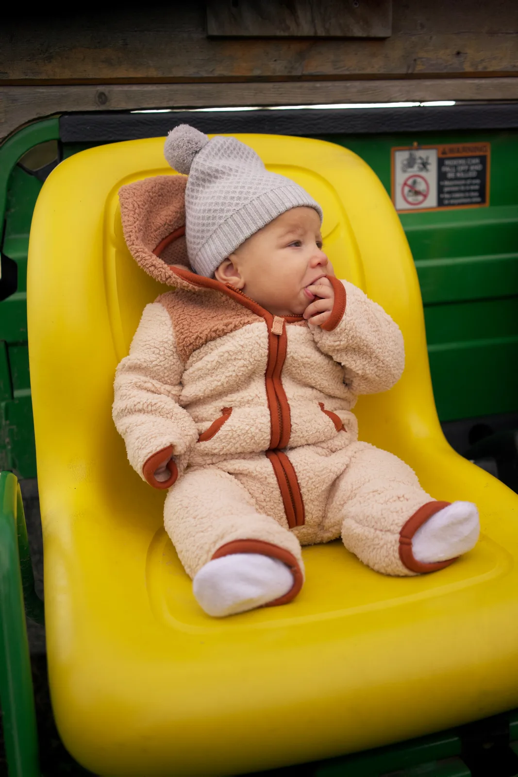 Baby in cream bear suit and gray knit hat seated on yellow tractor seat