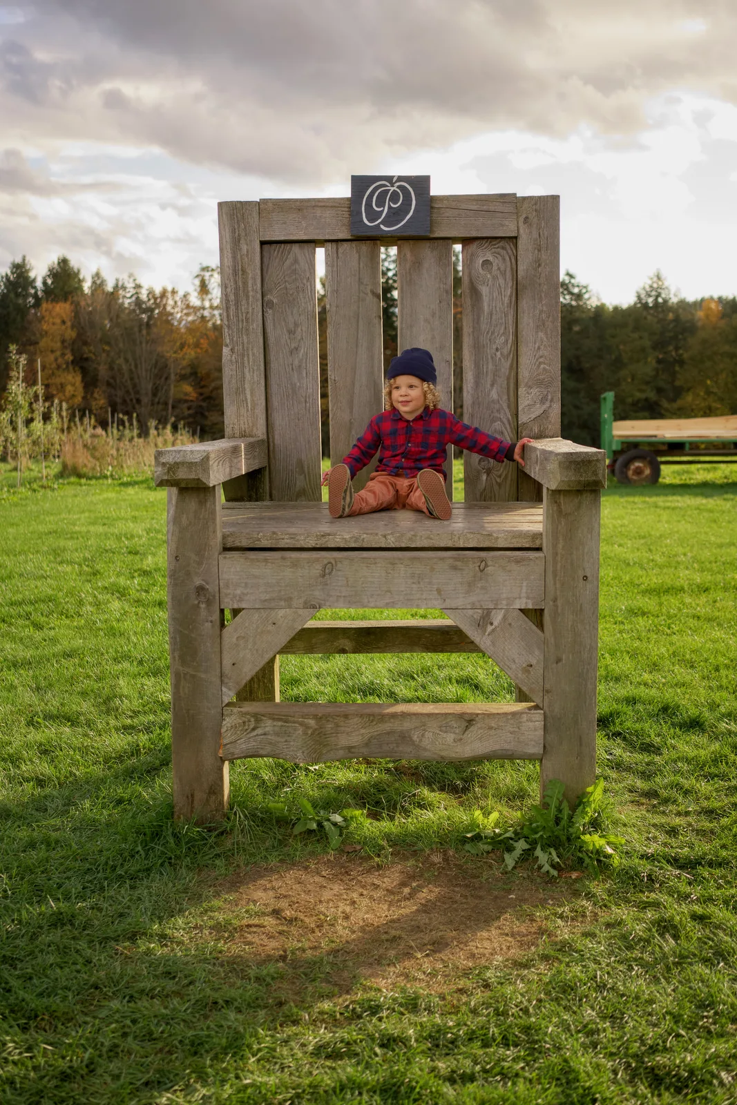 Child in red plaid shirt and navy beanie kneeling on large wooden chair, orchard and tractor in background