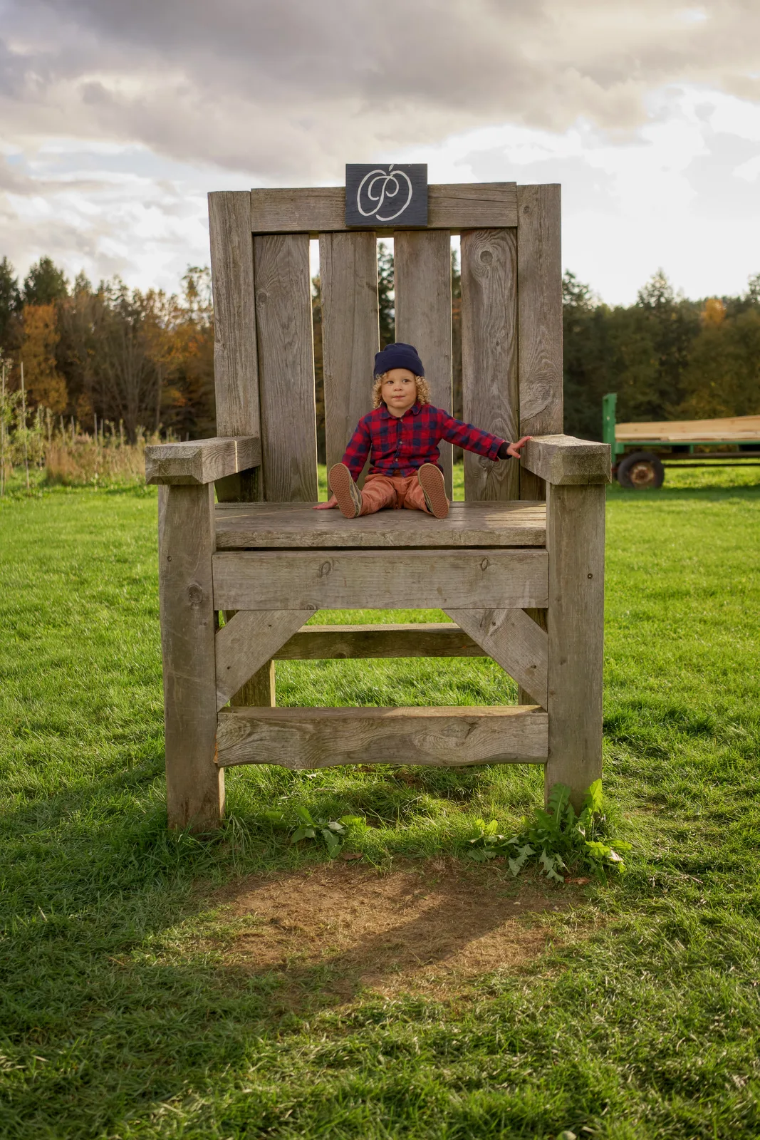 Child in red plaid shirt and navy beanie sitting on large wooden chair, green tractor and trees behind