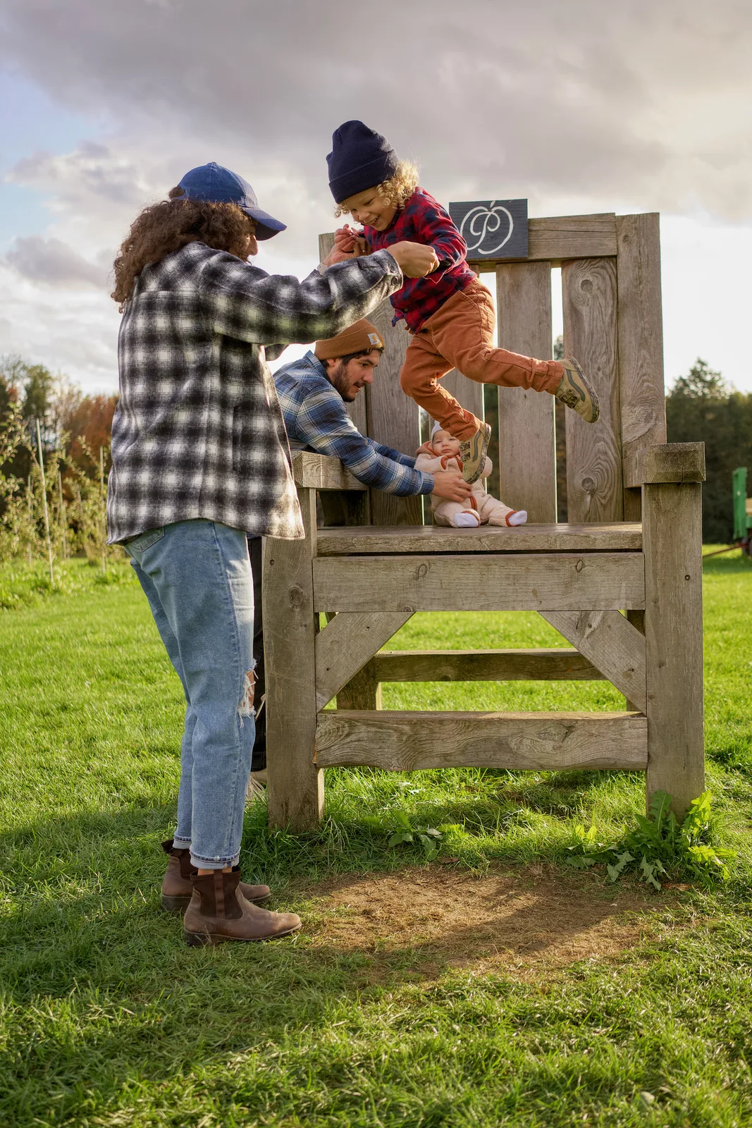 Two adults helping a child in red plaid shirt and blue beanie climb a large wooden chair