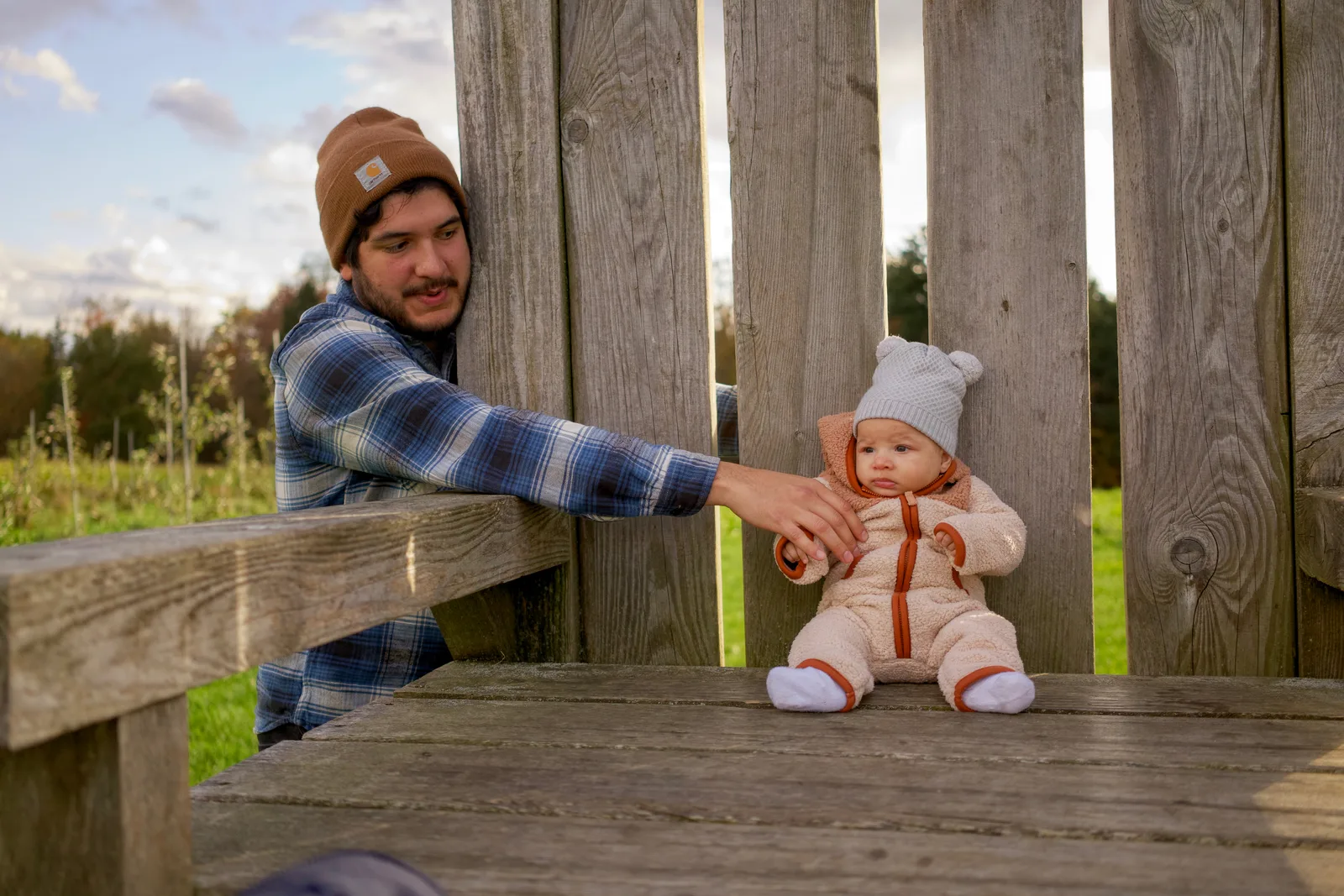 Adult in brown beanie and plaid shirt reaching for baby in cream bear suit on wooden bench