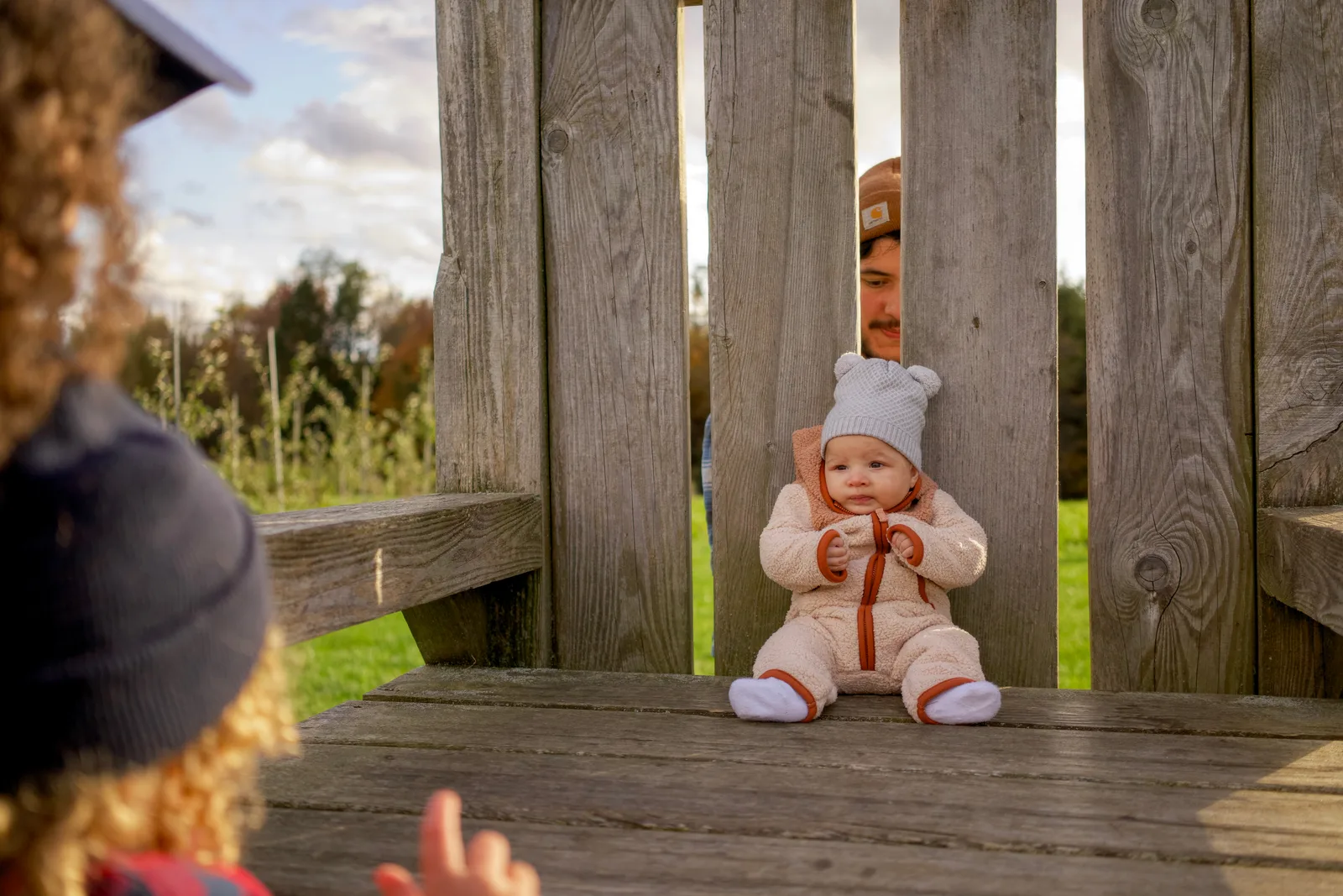 Baby in cream bear suit and white knit hat sitting on wooden bench between adults
