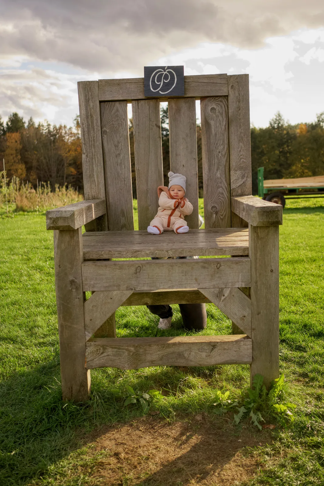 Baby in cream bear suit and white knit hat seated on large wooden chair in grassy field