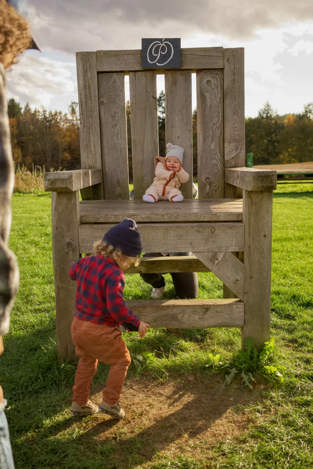 Child in red plaid shirt and blue beanie approaching large wooden chair with baby seated