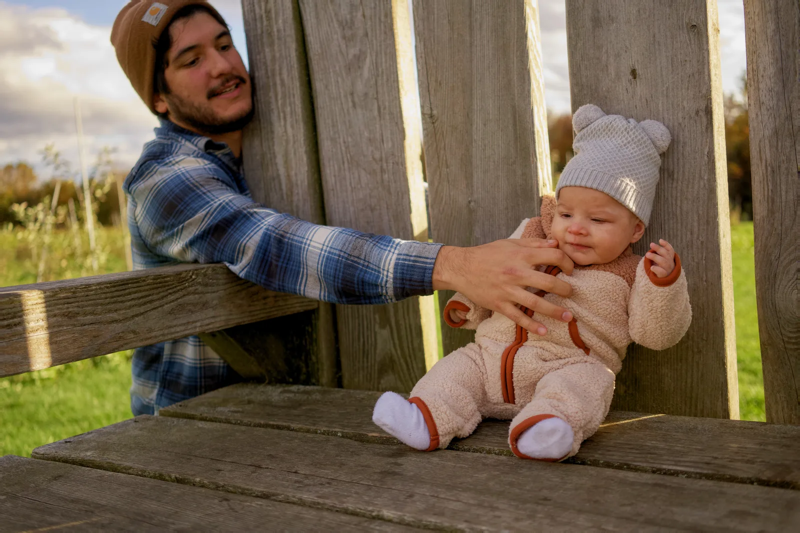 Adult in brown beanie and plaid shirt holding baby in cream bear suit on wooden bench