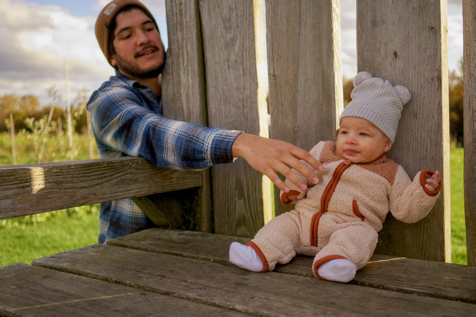 Adult in brown beanie and plaid shirt supporting baby in cream bear suit on wooden bench
