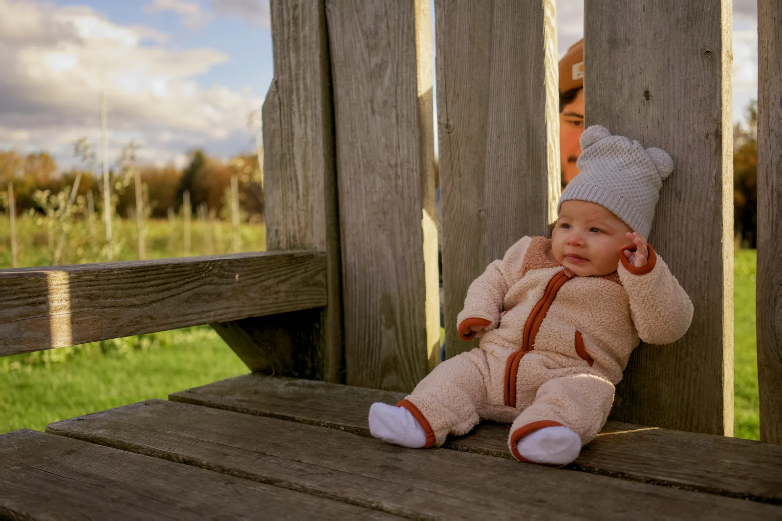 Baby in cream bear suit and white knit hat sitting on wooden bench between adults