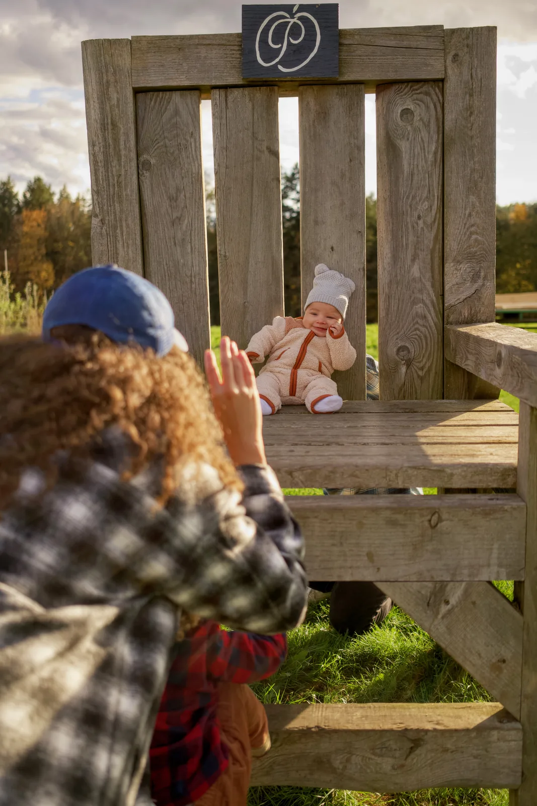 Adult in blue cap waving at baby in cream bear suit on large wooden chair