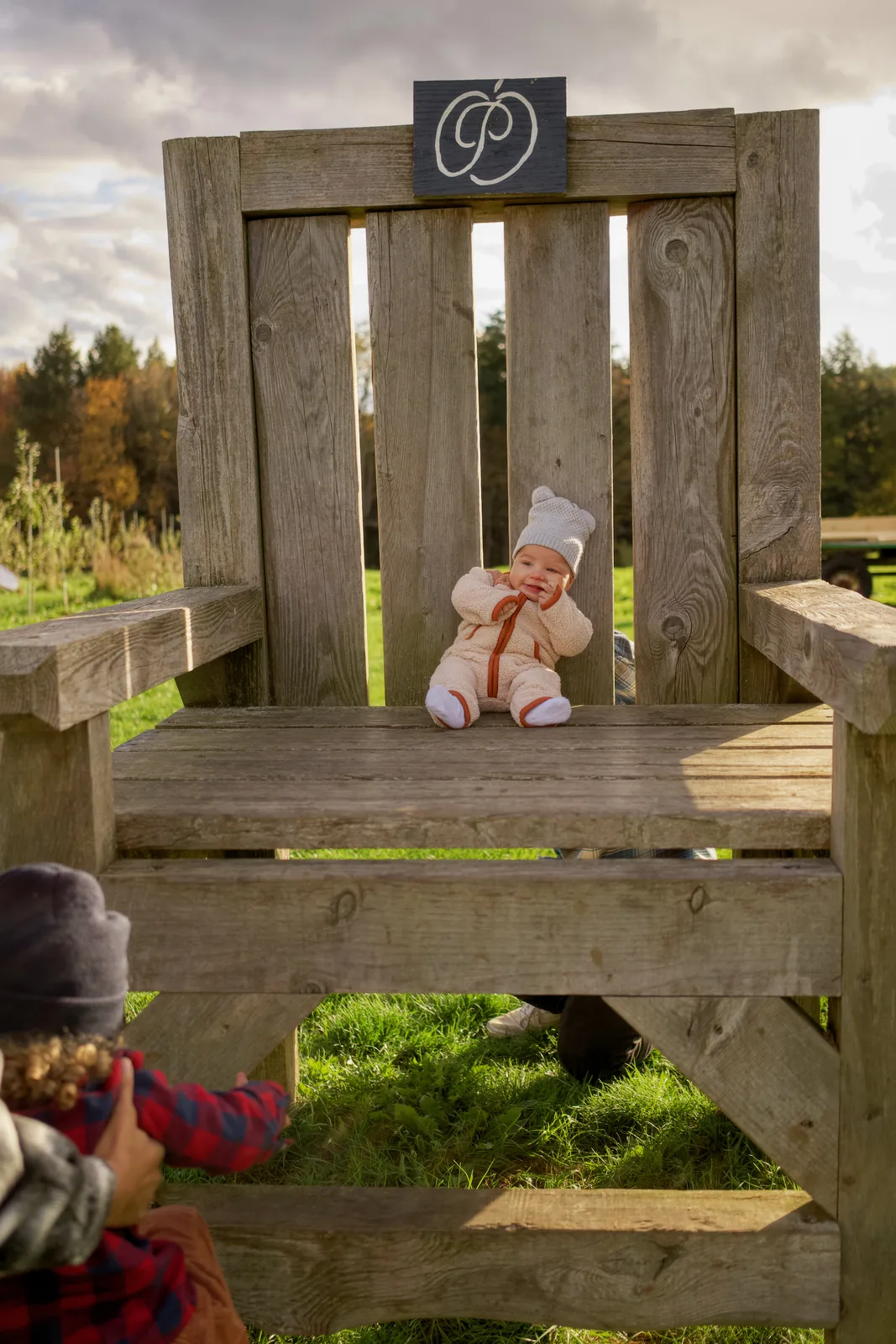 Child in gray beanie taking a photo of baby in cream bear suit on large wooden chair