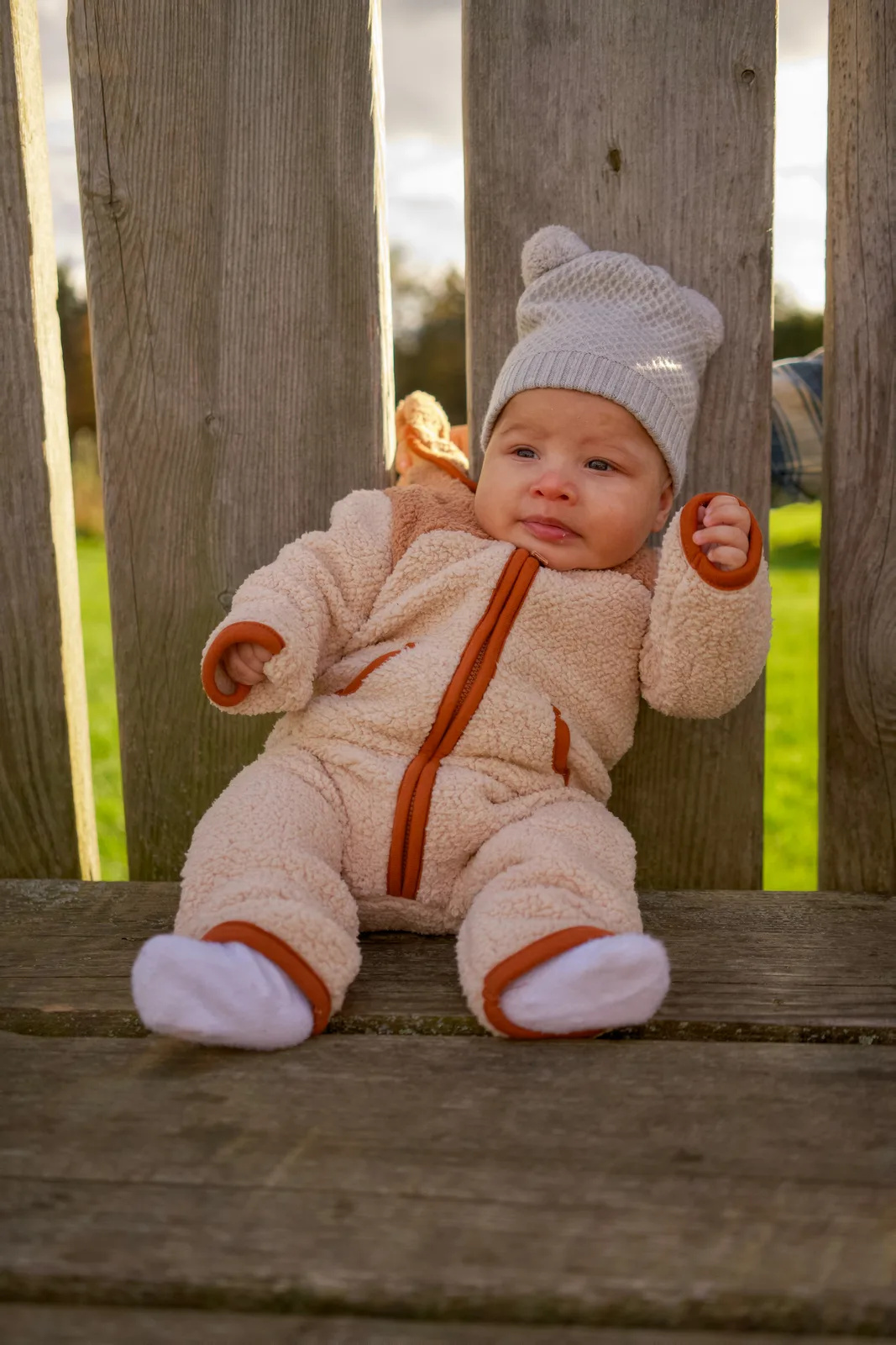Baby in cream bear suit and gray knit hat sitting on wooden bench