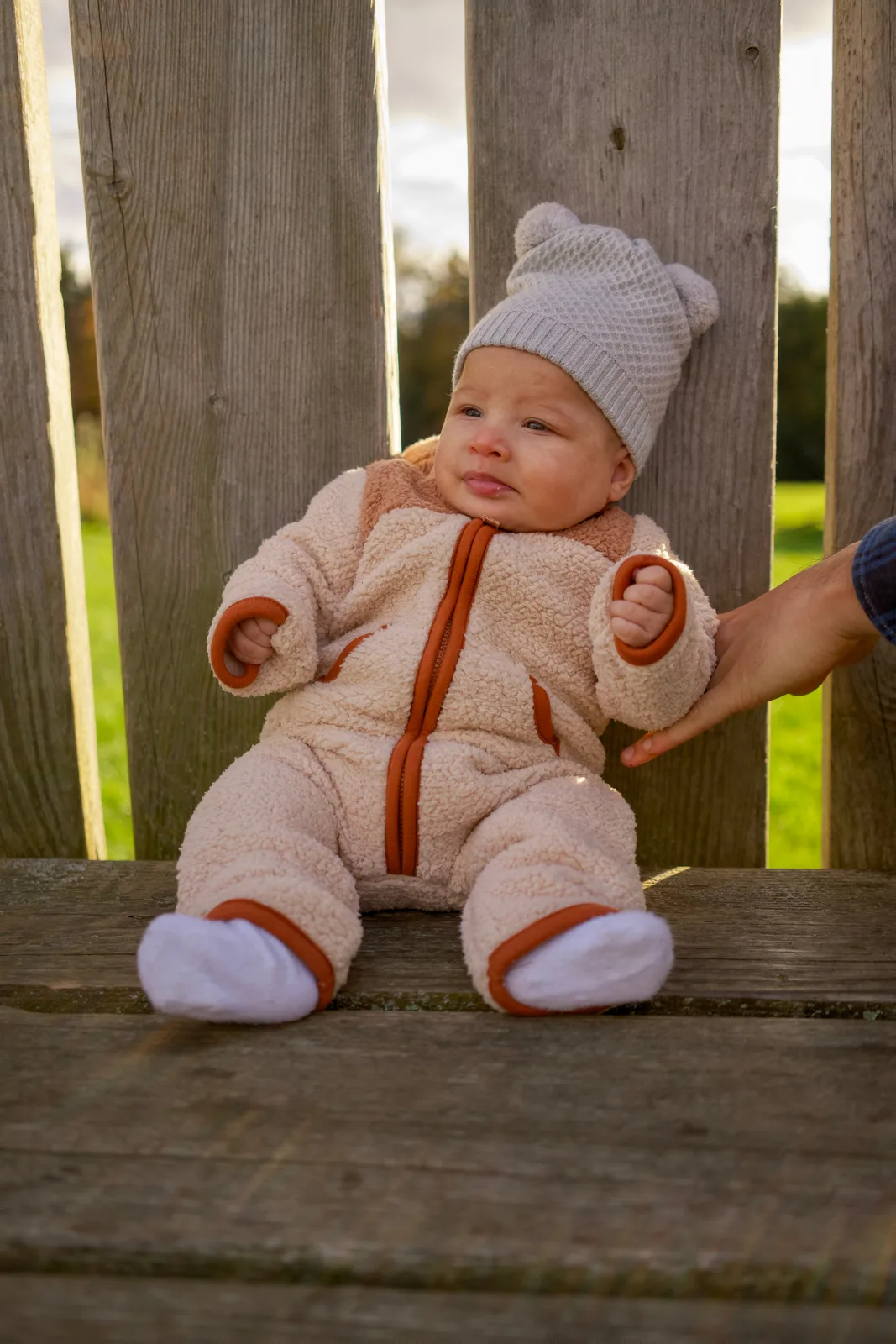 Baby in cream bear suit and gray knit hat on wooden bench, adult hand supporting