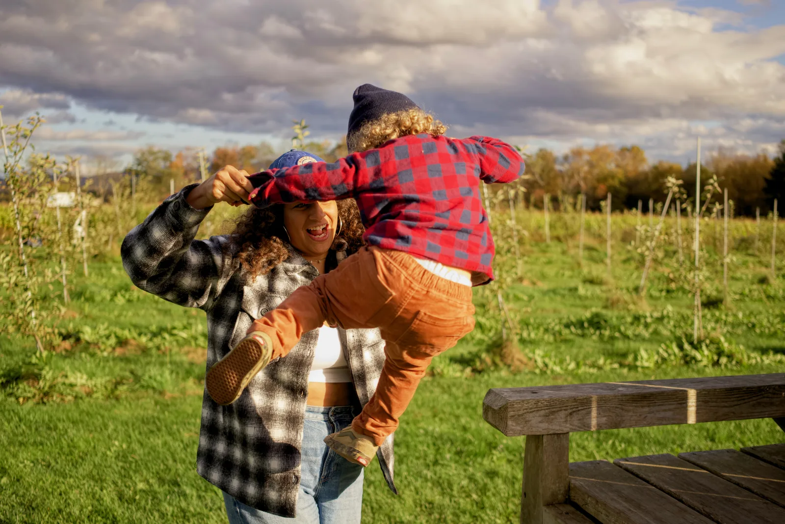 Adult in plaid shirt lifting child in red plaid shirt and blue beanie in grassy field