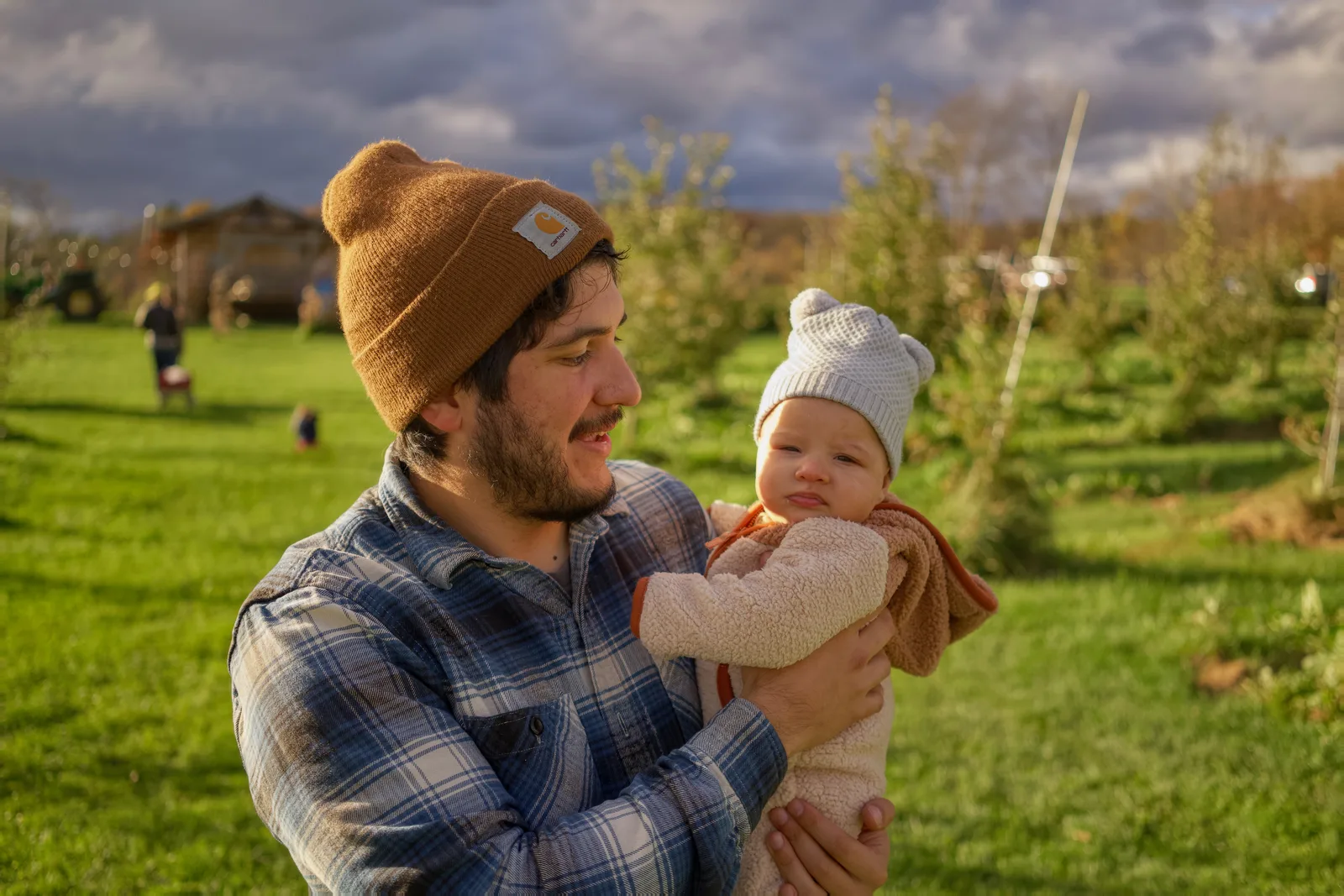 Adult in brown beanie and plaid shirt holding baby in cream bear suit in orchard