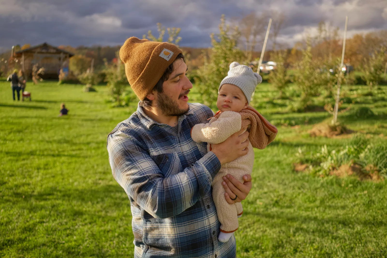 Adult in brown beanie and plaid shirt holding baby in cream bear suit, orchard rows behind