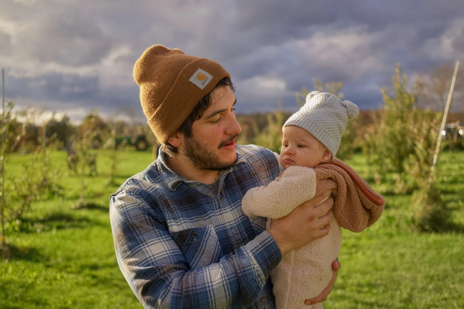 Adult in brown beanie and plaid shirt holding baby in cream bear suit, orchard rows behind