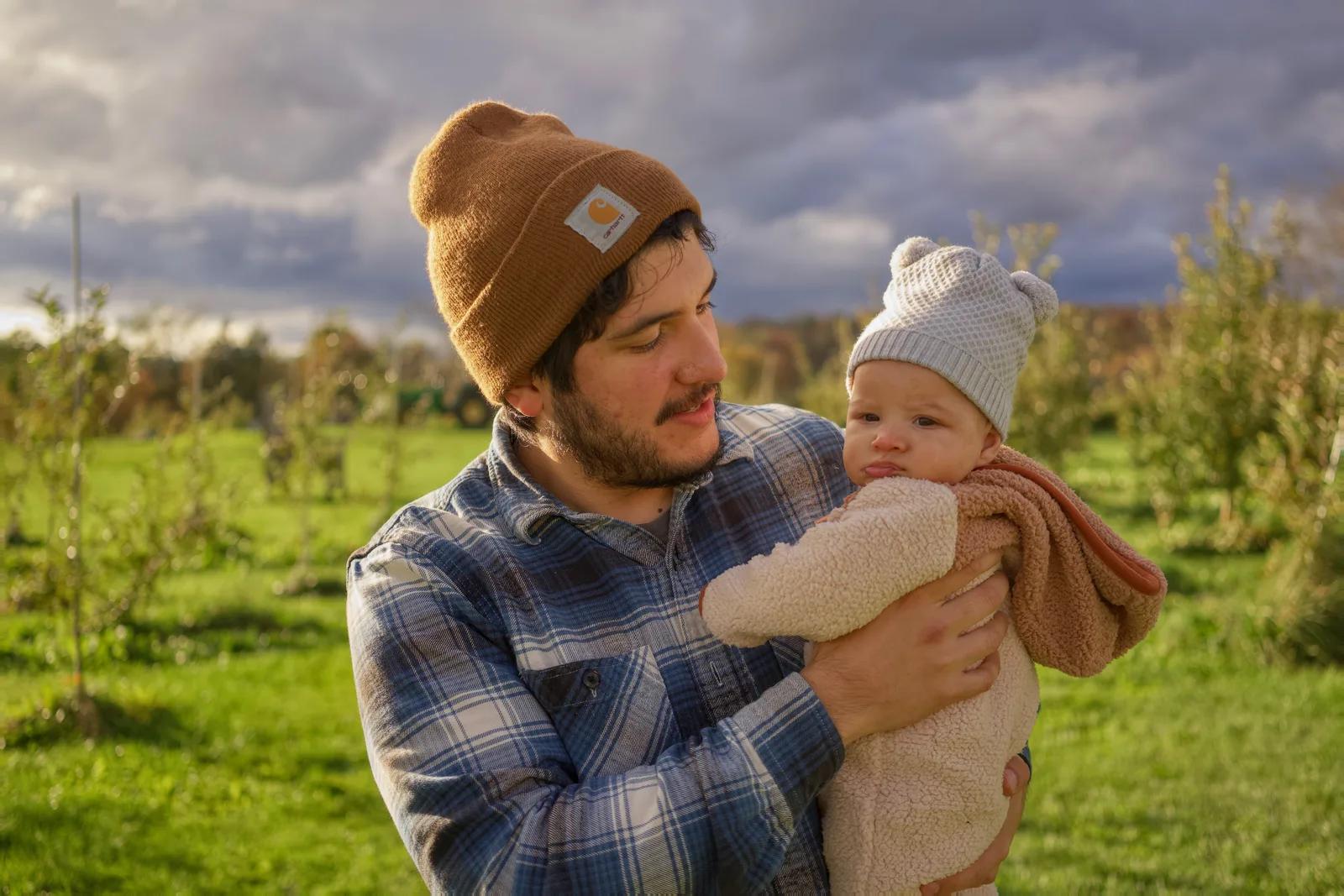 Adult in brown beanie and plaid shirt holding baby in cream bear suit, cloudy sky above