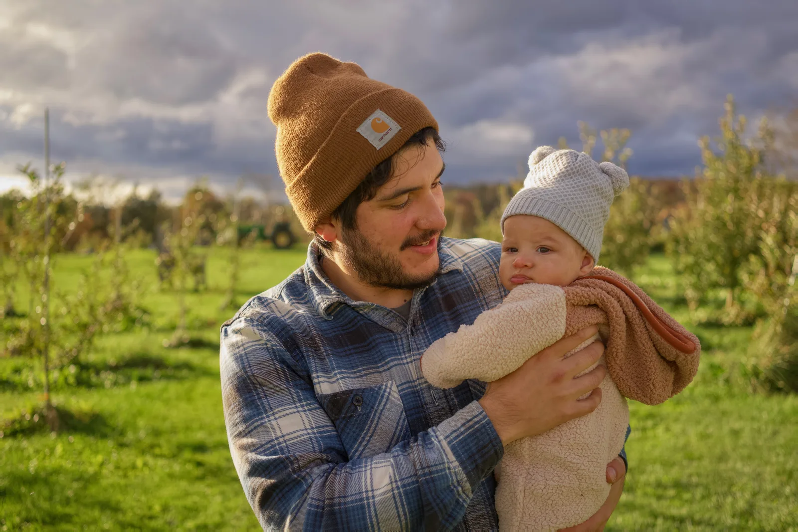 Adult in brown beanie and plaid shirt holding baby in cream bear suit, green field and trees