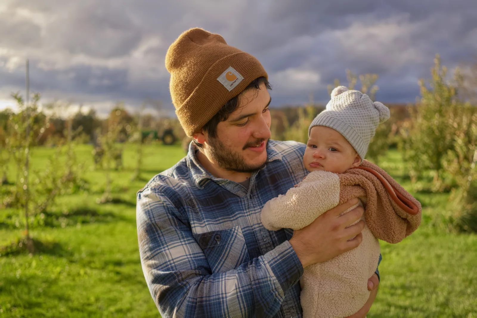 Adult in brown beanie and plaid shirt holding baby in cream bear suit, orchard and distant tractor