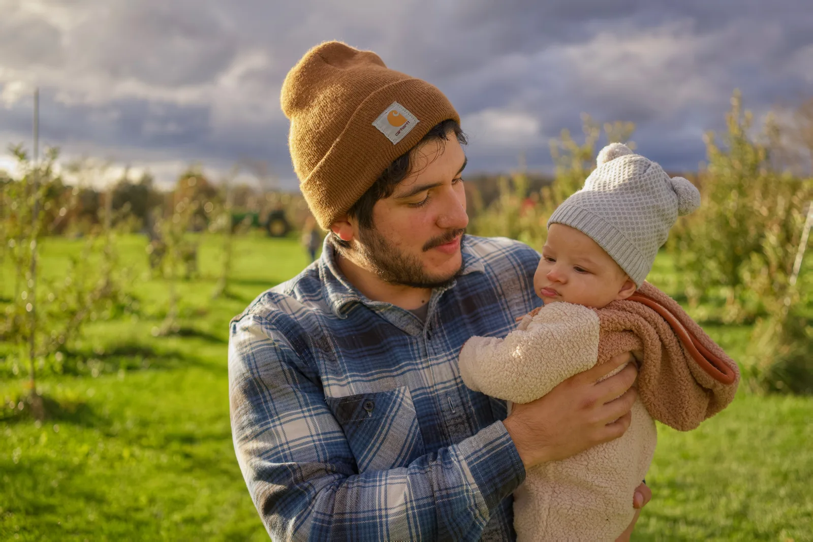 Adult in brown beanie and plaid shirt holding baby in cream bear suit, grassy orchard with trees