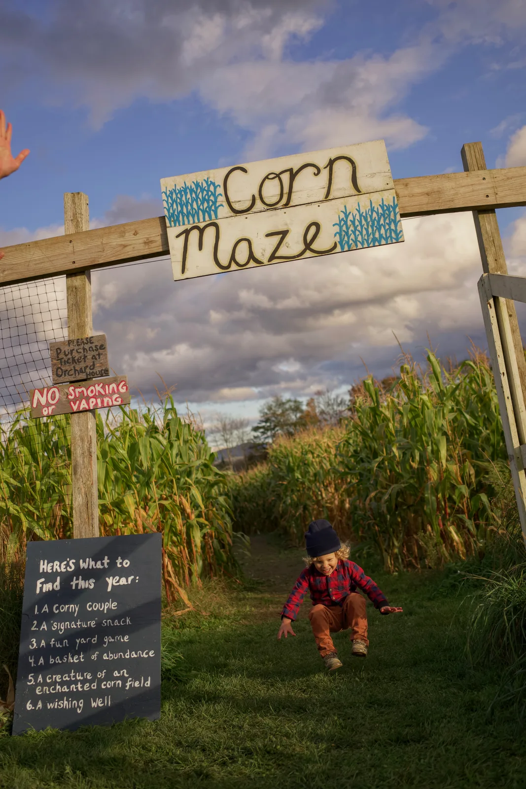 Child in red plaid shirt and blue beanie crawling under Corn Maze sign