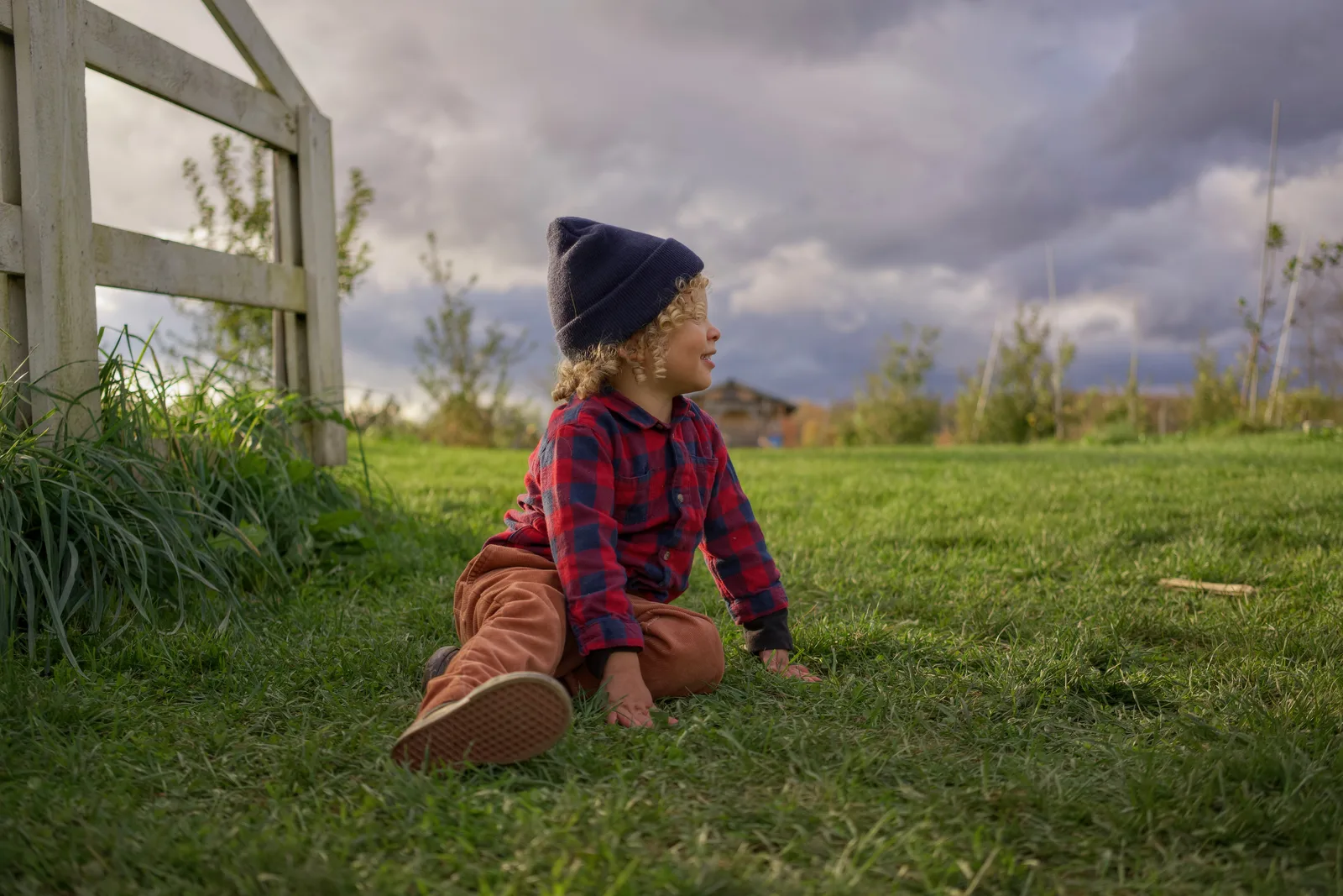 Child in red plaid shirt and blue beanie squatting near wooden fence in grassy field