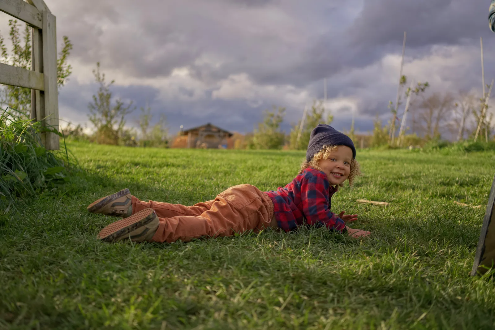 Child in red plaid shirt and blue beanie lying on grass near wooden fence